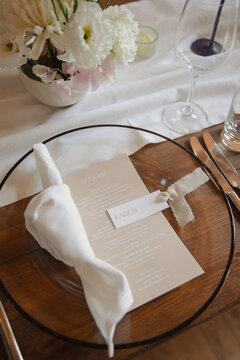 A beautifully set table with a clear glass plate, folded napkin, printed menu, and fresh floral arrangement, creating a refined and elegant atmosphere for a modern wedding table
