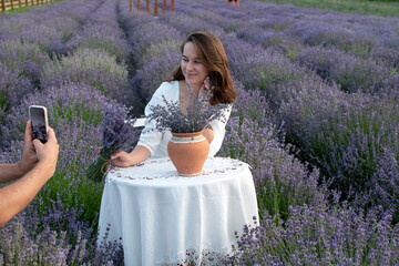 A man takes a photo of a woman in a white dress sits at a table in the middle of a lavender field	