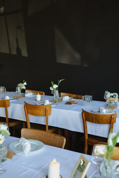A cozy and minimalist dining room setup featuring wooden chairs and white tablecloths, illuminated by soft natural light against a dark wall background. The table is adorned with simple floral arrange