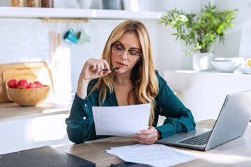 Pretty young business woman working with computer while consulting some invoices and documents in...