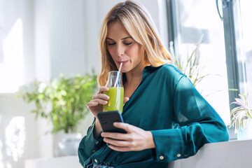 Beautiful woman using her mobile phone while drinking fruit detox juice in the kitchen at home.