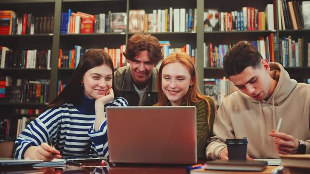 Group of students collaborating at table in library, working together on laptop and notebooks. Bookshelves filled with colorful books in background. Concept of teamwork and academic collaboration