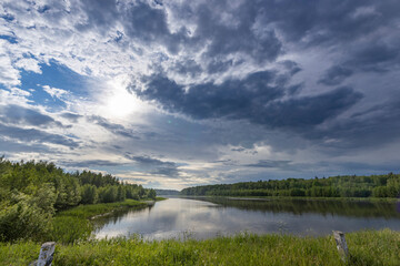 Obraz premium Tranquil river with lush trees, dramatic sky and sun peeking through swirling clouds, creating a picturesque moment in nature.