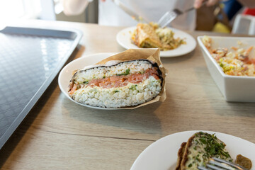A close-up image of a delicious sushi burrito filled with salmon and rice on a white plate. The burrito is partially unwrapped from its paper wrapping