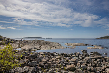 Weathered rocks dot the shoreline with islands in the distance and a reflective pool of clear water mirroring the blue sky and clouds above.