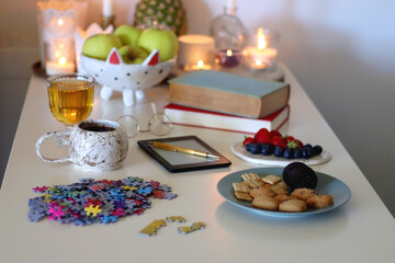 Colorful jigsaw puzzle pieces, books, tablet, glasses, cookies, berries, cup of tea or coffee, glass of juice and lit candles on the table. Hygge in pastel colors. Selective focus.