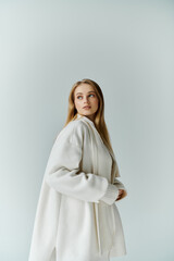 A young woman in a white coat stands against a light blue wall, looking to her right.
