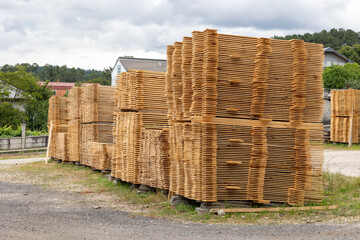Large stacks of neatly arranged wooden boards are stacked outdoors on a bright, sunny day, likely for construction or woodworking purposes, against a natural backdrop.