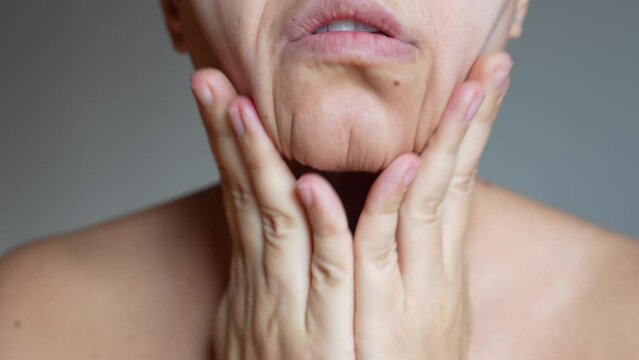 Cropped shot of young caucasian woman massaging her jaw grabbing with effort the skin of lower face on a dark grey background. Rejuvenation, facelift, facefitness, skin care. Cosmetology and beauty