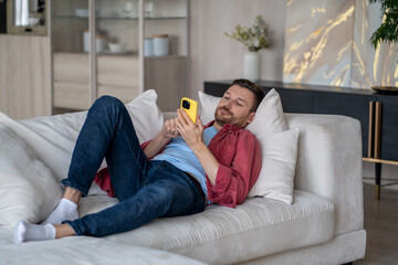 Lazy man lies on sofa in living room, uses phone, thoughtlessly meaninglessly scrolling through feed of social networks, absorbing information, content. Tired calm guy resting on couch with smartphone