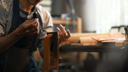 Professional craftsman in apron sewing while making handmade leather wallet in workshop