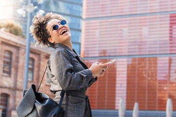 Cute African girl with ponytail standing on the street and using smartphone on modern buildings background.