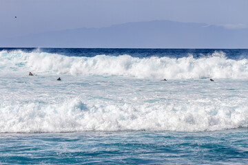 Waves coming ashore on a sunny day and surfers ready to surf the waves. Wall of blue wave with white foam coming ashore where surfers are surfing. Atlantic Ocean. Tenerife, Canary Islands, Spain.