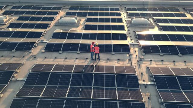 Aerial shot of engineers in helmets walking across photovoltaic array on rooftop. Electricians inspecting solar panels electrical safety at sunset rotating drone shot