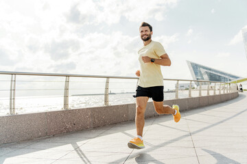 A man jogging on a waterfront promenade on a sunny day, wearing a yellow shirt and black shorts, with modern buildings in the background.