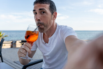 Man taking selfie while drinking spritz cocktail by the sea. Man enjoying a refreshing spritz cocktail while taking a selfie on a sunny day at a beach bar