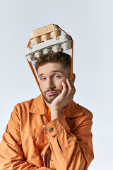 A man in an orange jacket wears an egg carton on his head.