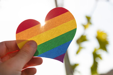 Hand holding a paper heart in LGBT rainbow colors with the sun rays shining around it, concept of...