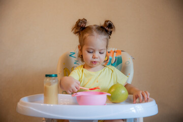 A little girl is sitting on a high chair and eating mashed fruit at home