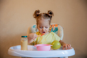 A little girl is sitting on a high chair and eating mashed fruit at home