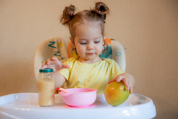 A little girl is sitting on a high chair and eating mashed fruit at home