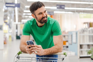 Smiling bearded man looking away holding smartphone and leaning on the shopping cart