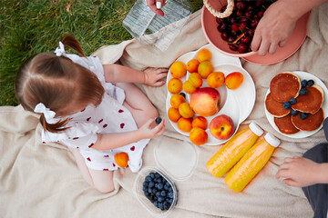 A baby with blonde hair on a picnic with her mother and brother eating fresh fruits, pancakes