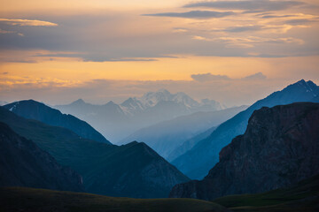 Deep gorge between layered silhouettes of sharp rocky ridge spurs under huge snowy mountain peak in golden cloudy sky. Sheer crags of mount valley under giant ice top under gold sunset color clouds.