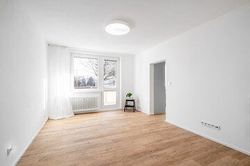 A bright, empty living room with white walls, wooden parquet flooring, a large window with white curtains, a radiator, and a small black stool with a plant.