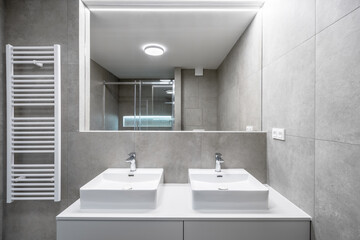 A modern bathroom with gray cement tiles, a double sink with chrome faucets, a large mirror, a glass-enclosed shower, a wall-mounted towel heater, and LED strip lighting.