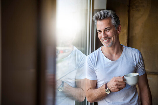 Smiling man holding coffee cup and leaning on glass window at cafe - Powered by Adobe