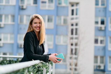 Fototapeta premium Blonde Businesswoman in Black Suit Using Smartphone on Break in Modern Urban Environment.