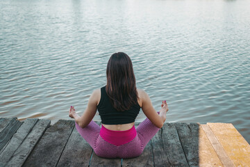 Back view, young sporty woman practicing meditation in lotus pose on wooden pier by a river, morning workout outdoors