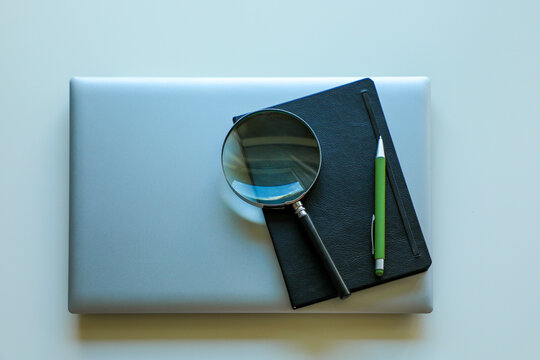 On top of the laptop there is a notepad, a green pen and a magnifying glass, close-up. - Powered by Adobe