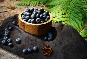 Blueberries in a wooden board bowl on a black linen napkin on a wooden table surrounded by green fern