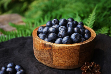 Blueberries in a wooden board bowl on a black linen napkin on a wooden table surrounded by green fern