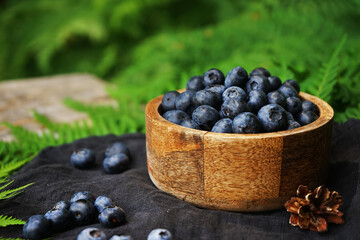 Blueberries in a wooden board bowl on a black linen napkin on a wooden table surrounded by green fern