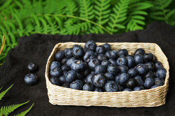 Blueberries in a wicker bowl on a black linen napkin on a wooden table surrounded by green fern