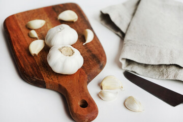 Whole garlic and cloves on a wooden board next to a knife and linen napkin on a light background