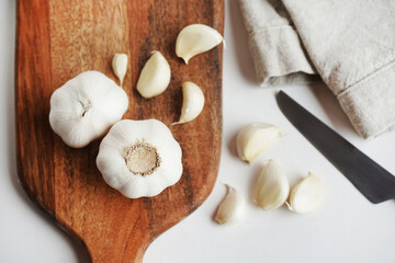 Whole garlic and cloves on a wooden board next to a knife and linen napkin on a light background