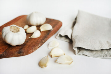 Whole garlic and cloves on a wooden board next to a knife and linen napkin on a light background