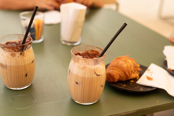 Two glasses of iced coffee with a croissant on a green table