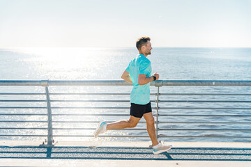 Jogger running along a sunlit seaside boardwalk, ocean sparkling under the bright morning sun.