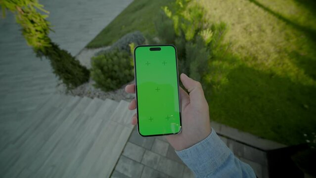 Man hands holding and scrolling vertically phone with green screen. On background stairs outdoors. Nature grass