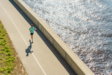 Aerial view of a jogger running along a sunlit riverside path, casting a shadow beside the...