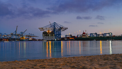 Bustling port scene at night featuring cargo ships, illuminated cranes, and their reflections on calm water, viewed from sandy shoreline