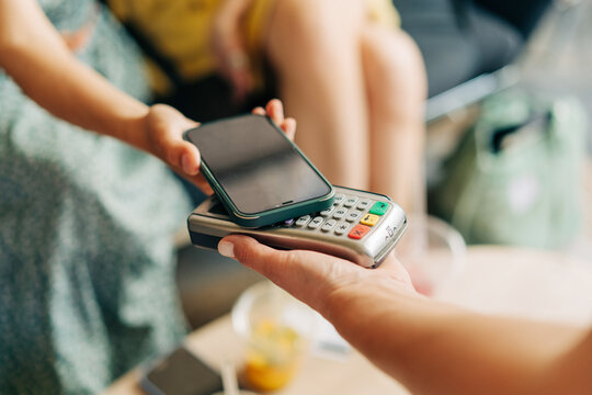 Close up view at hand hold mobile phone, touch a credit card reader inside the coffee shop. Cashless payment concept.
