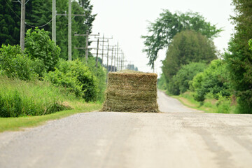 A large round bale of hay from an insecure load blocking a country road that has fallen off a hay wagon 
