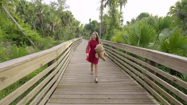 Teenage girl walking alone with her teddy bear on wooden boardwalk trail through wild tropical jungles. Child exploring Florida nature in dense green rainforest