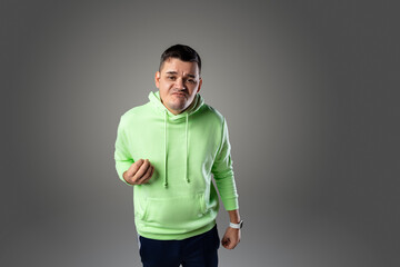 A young man wearing a green hoodie stands against a gray backdrop. He has a serious expression on his face and is clenching his fist.